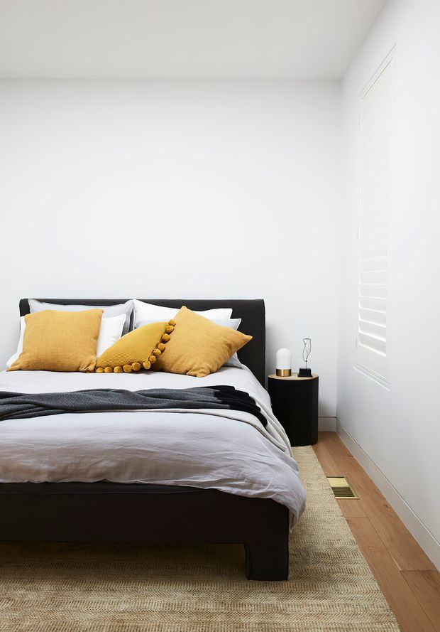A bright, minimalist bedroom with a black upholstered bed, crisp white bedding, mustard yellow cushions, and a dark charcoal throw. A small black round bedside table, white plantation shutters, pale timber flooring, and a neutral textured rug complete the clean, contemporary space.