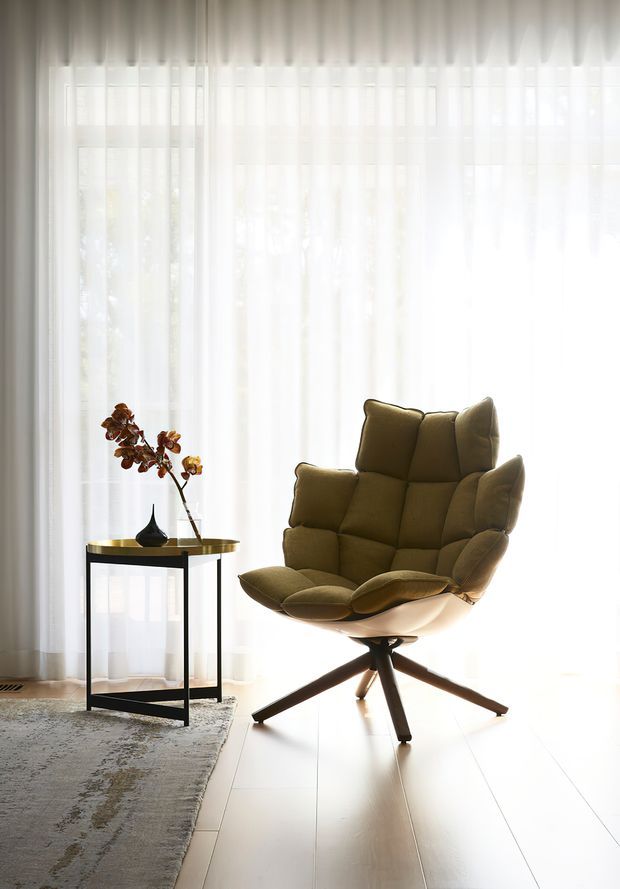 A bright, minimalist sitting area with sheer floor-to-ceiling curtains, pale timber flooring, and a sculptural olive-brown upholstered lounge chair on a star-shaped wooden base. A slim black metal-and-glass side table with a small vase sits beside the chair, while a muted patterned rug occupies the foreground.