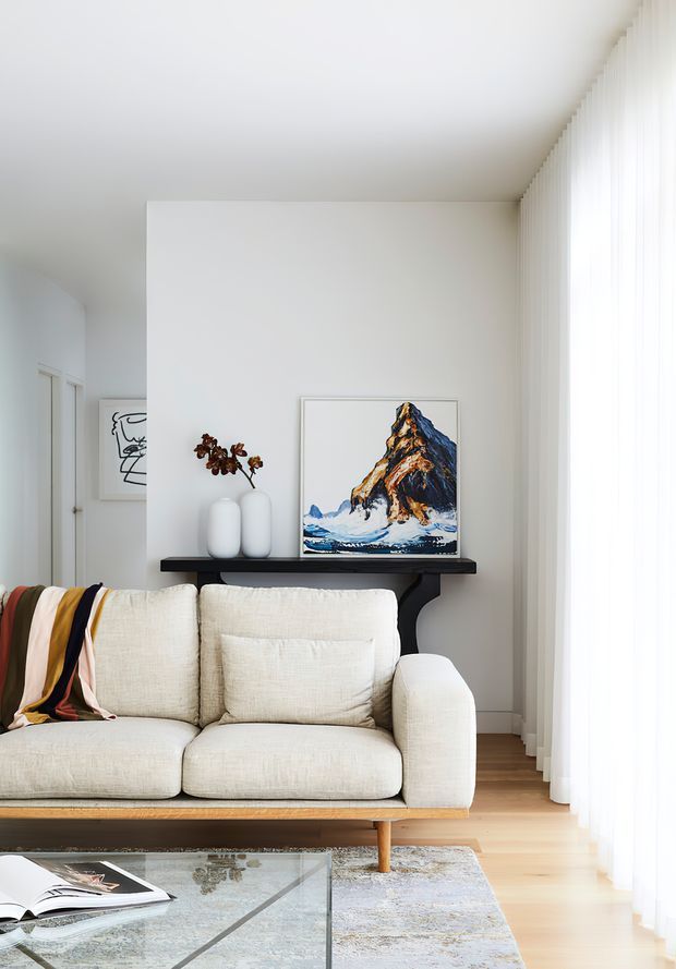 Bright, calm living room with a cream upholstered sofa, pale timber flooring, sheer floor-to-ceiling curtains, a black console table, ceramic vases, and mountain artwork. A glass coffee table and patterned rug sit in the foreground, with strong natural light entering from the right.