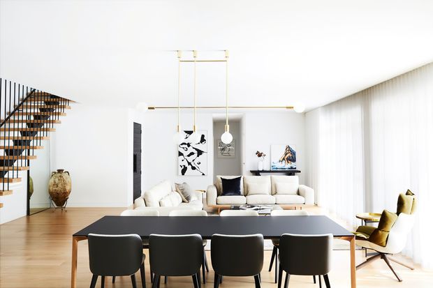 Bright, open-plan contemporary living and dining space with a large dark dining table and upholstered dining chairs in the foreground, a light sectional sofa and lounge chairs beyond, timber flooring, expansive sheer-curtained glazing, and a floating open-riser staircase with black railings. Minimal decorative styling and sculptural pendant lighting create an airy, refined interior.