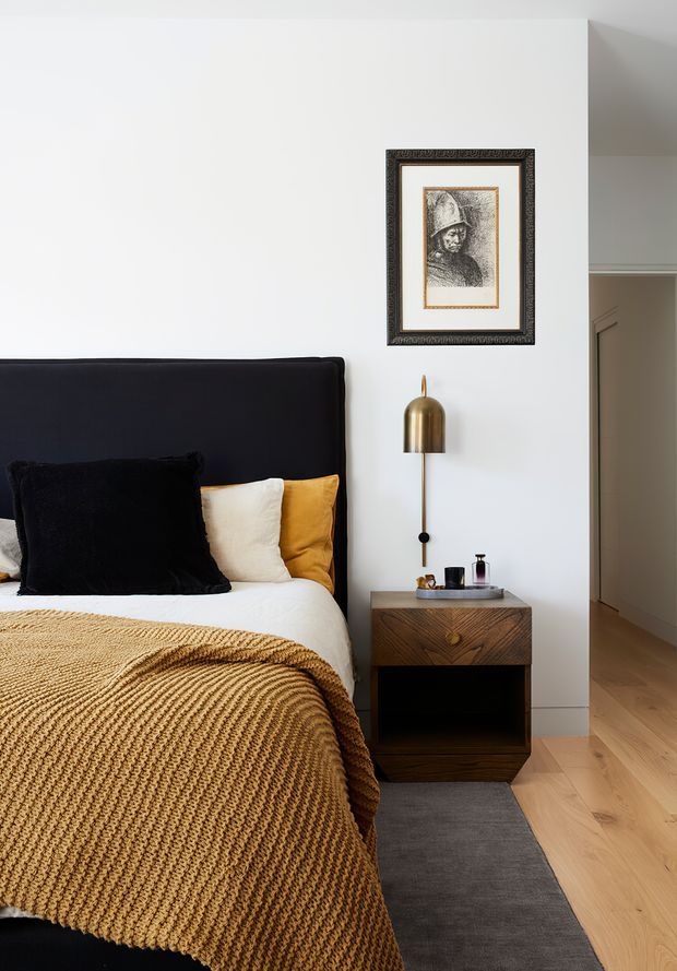 A refined bedroom interior featuring a dark upholstered bedhead, white bedding, black and cream cushions, and a mustard knitted throw. A compact dark timber bedside table with brass detailing sits beneath a sculptural brass wall lamp. White walls, pale timber flooring, a grey rug, and framed artwork create a warm, elegant scheme. A hallway or adjoining space is visible through the opening at right.