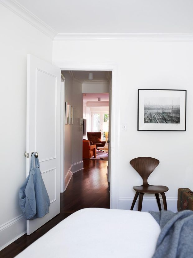 Bright, refined bedroom viewed toward an open doorway leading to a warmly furnished sitting room. The room has white walls and ceiling with decorative crown moulding and recessed wall panelling, dark timber flooring, a white upholstered bed in the foreground, a sculptural dark wooden chair, framed monochrome artwork, and a paneled white door with a hanging blue garment. The adjoining room contains a rust-coloured lounge chair and additional traditional detailing.