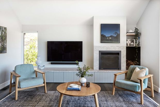A bright, compact living room with a contemporary fireplace wall, wall-mounted television, low built-in media cabinetry, and two light blue upholstered timber-framed armchairs facing a round wooden coffee table. The room has white walls and ceiling, dark timber flooring, a large textured area rug, minimalist shelving, and a window providing natural light.