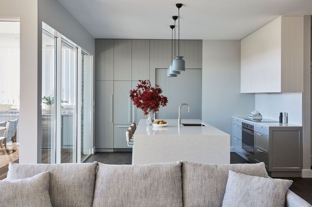 A bright contemporary open-plan kitchen viewed from behind a light neutral sofa. The space features a large pale stone island with an undermount sink and curved faucet, full-height grey cabinetry, integrated appliances, white upper cabinetry, dark timber flooring, black-framed glass doors, and three suspended grey pendant lights. A dining area is visible through the glazed opening on the left.