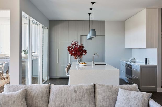 Contemporary open-plan kitchen viewed from the living area, featuring a large pale stone island with integrated sink, full-height grey cabinetry, dark flooring, white walls, expansive glazed doors, and three pendant lights. A sofa occupies the foreground, with a dining area visible beyond the glazing.