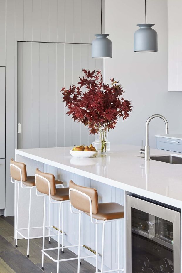 Bright, refined contemporary kitchen interior featuring a large white island with integrated sink, brushed metal tapware, white vertical-panel cabinetry, a built-in wine refrigerator, and three tan leather-and-white bar stools. Grey pendant lights hang above the island, while burgundy foliage in a glass vase provides a natural focal point. Dark timber flooring adds contrast.