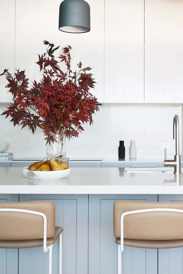 A bright, refined contemporary kitchen viewed across a white island bench. Pale blue-grey panelled cabinetry and matching upper cupboards create a clean backdrop, with a stainless-steel gooseneck tap, minimalist black and white vessels, and a large charcoal pendant light. Two tan upholstered bar stools sit at the island, while a glass vase of burgundy foliage and a bowl of pears provide natural colour accents.