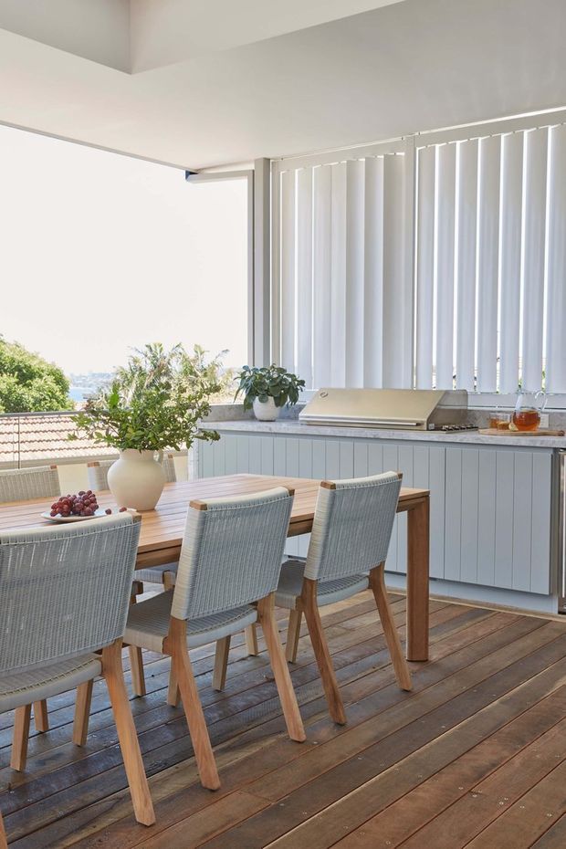 A bright covered outdoor dining and kitchen area on a timber deck. A long timber dining table with woven light-grey chairs faces a built-in pale blue-grey cabinet run and countertop with a barbecue. White vertical blinds enclose the space, while greenery and a distant coastal or urban view are visible beyond the opening. The overall look is relaxed, light, and coastal.