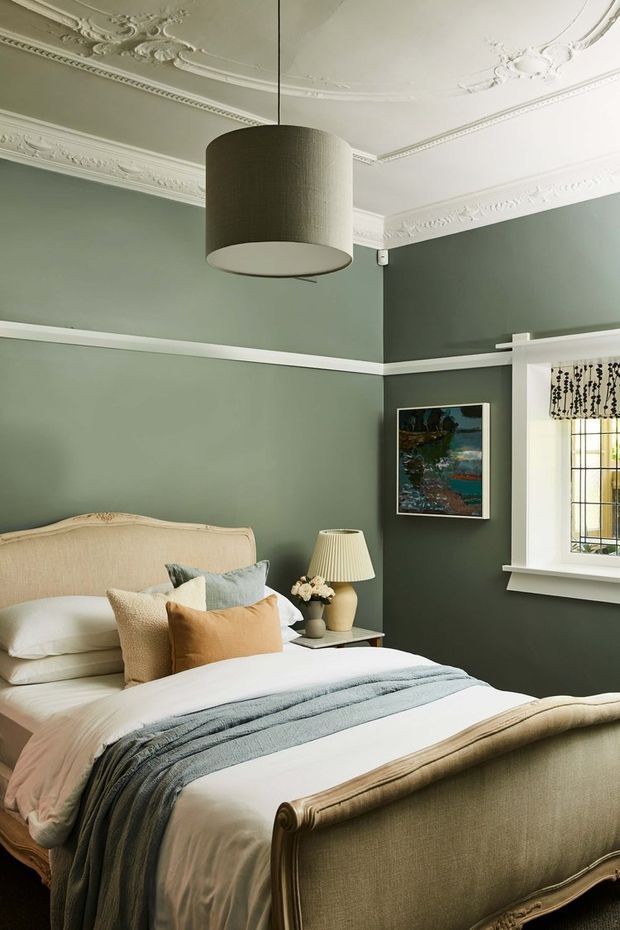 A refined bedroom with deep muted green walls, ornate white ceiling cornices and plaster detailing, and a classic upholstered bed with curved carved-style end panels. Layered cream, pale blue, and ochre cushions and bedding create a warm, elegant look. A large cylindrical pendant hangs centrally, while a bedside table, lamp, artwork, and decorative leadlight-style window complete the room.