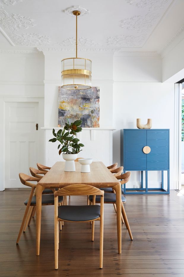 Bright, refined dining room with a timber dining table and matching curved wooden chairs, set against dark timber flooring and white decorative wall panelling. A blue built-in-style sideboard with a brass-toned circular handle adds a bold accent, while a large brass and glass pendant hangs from an ornate white ceiling with decorative plaster detailing. Artwork, a potted plant, and sculptural tableware complete the space.