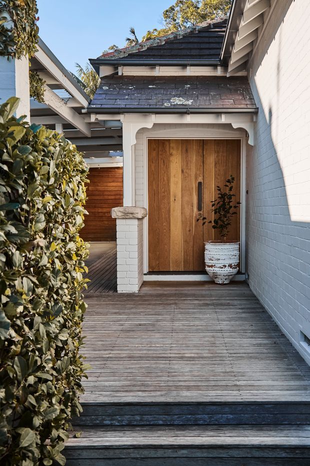A covered exterior entry approached by a weathered timber deck, with a warm vertical timber front door set into a white-painted brick and rendered surround. The entry has decorative scalloped trim, a dark shingle-clad pitched roof, exposed eaves, and dense hedging along the walkway. A white distressed planter sits beside the door, creating a welcoming coastal-traditional character.