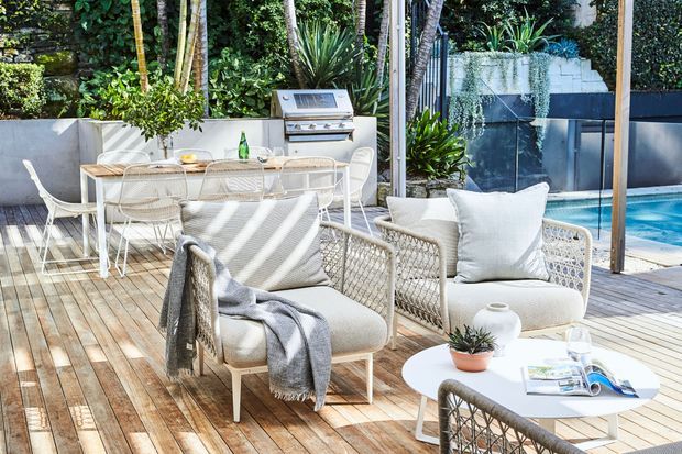 A bright outdoor entertaining area on a timber deck, furnished with woven lounge chairs, a low round coffee table, and a dining table with chairs. A built-in stainless-steel barbecue sits against a rendered wall, with a swimming pool and lush planting beyond. The space has a relaxed contemporary resort-like feel with pale upholstery, natural timber, and abundant greenery.