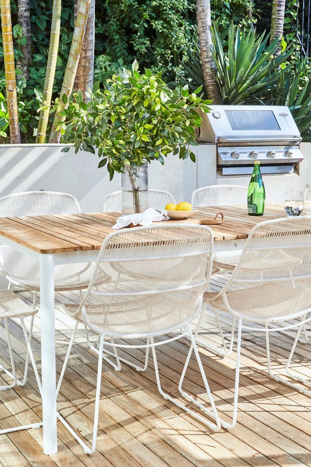 Bright outdoor dining area on a timber deck, featuring a large rectangular dining table with a timber slat top and white legs, woven white dining chairs, a built-in stainless-steel barbecue set against a low wall, and lush tropical planting behind it.