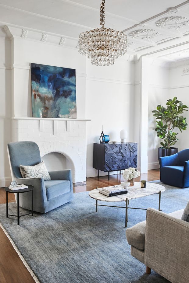 Elegant formal living room with white ornate mouldings, a decorative painted brick fireplace, large abstract artwork, crystal chandelier, blue velvet armchairs, patterned dark sideboard, marble coffee table, and a large blue-grey area rug over timber flooring.