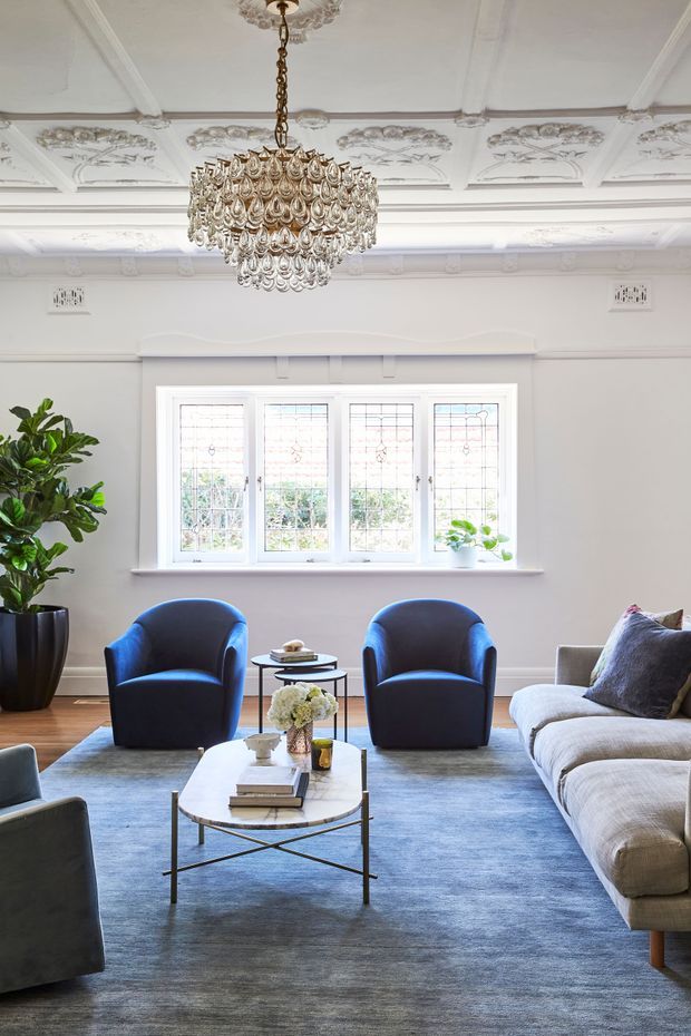 Elegant formal living room with ornate white decorative ceiling panels and mouldings, a large crystal chandelier, pale walls, stained-glass-style multi-pane windows, deep blue velvet armchairs, a light neutral sectional sofa, and a blue-grey area rug. The room combines heritage architectural detailing with refined contemporary furnishings.