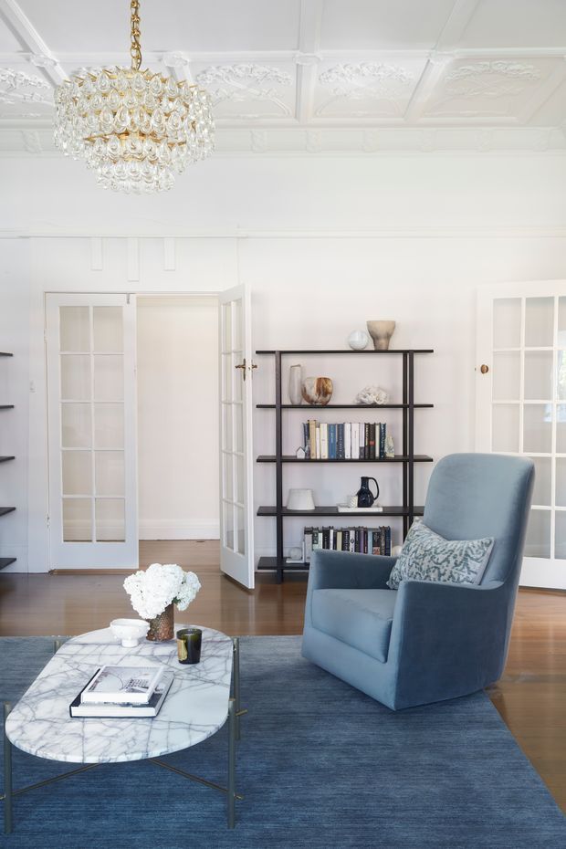 Bright, refined living room with a white decorative coffered ceiling, ornate plaster mouldings, a crystal chandelier, French doors, dark open shelving, and polished timber flooring. A blue upholstered armchair sits on a large blue rug beside a white marble oval coffee table, creating a classic yet contemporary interior.