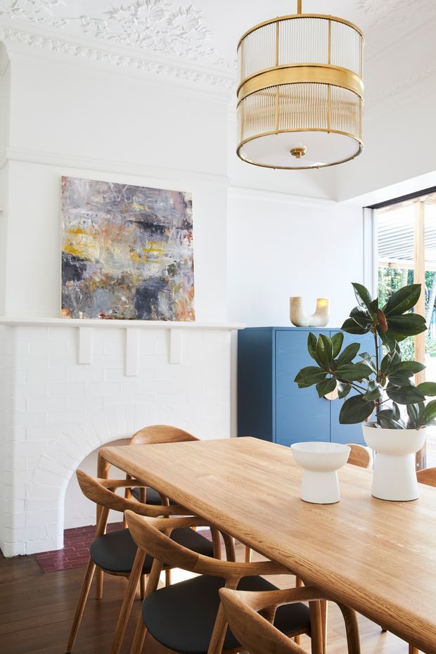 Bright, refined dining room with a large light-wood dining table and curved wooden chairs, centred around a white painted brick fireplace with an ornate decorative ceiling and cornice. A vivid blue built-in cabinet, abstract artwork, leafy indoor plant, and a large brass-and-ribbed-glass pendant add colour and character. Timber-framed glazing brings in abundant natural light.