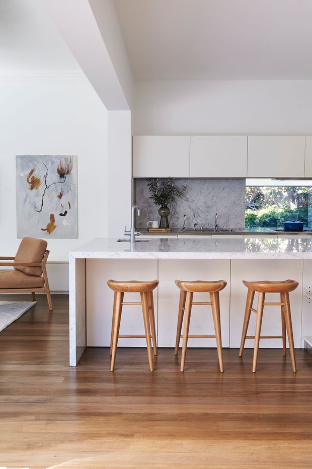 Bright contemporary kitchen with a large pale stone island and waterfall edge, white flat-panel cabinetry, a grey veined stone splashback, timber bar stools, and warm timber flooring. A sculptural abstract artwork and lounge chair are visible in the adjacent living area. Natural light enters through the kitchen window, creating an airy, refined interior.