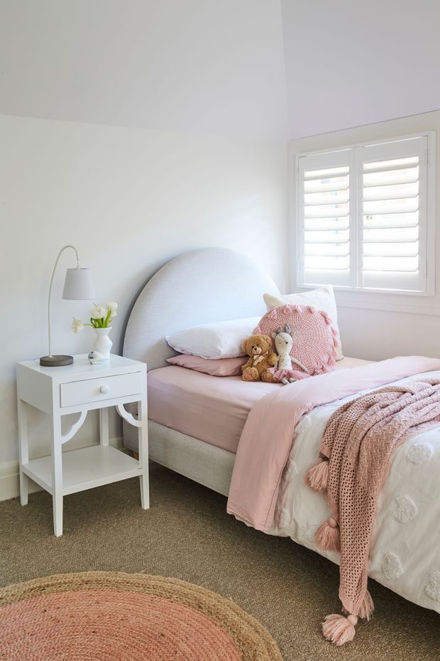A bright, softly styled child's bedroom with a single bed, rounded upholstered headboard, pink and white bedding, plush toys, a white bedside table with lamp and flowers, plantation shutters, and a neutral carpet. The palette is predominantly white, blush pink, and beige, creating a calm and playful atmosphere.
