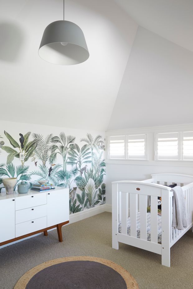 Bright nursery with white walls and ceiling, a grey pendant light, white plantation-shutter windows, and a white timber cot. A leafy tropical botanical wallpaper feature wall adds colour behind a mid-century-style white dresser with tapered timber legs. The room has neutral carpet and a round woven rug.