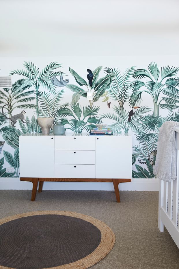 A bright, styled bedroom corner featuring a white mid-century-inspired sideboard with tapered timber legs, placed against a bold tropical botanical wallpaper. A white bed frame and textured throw are visible at right, with a round woven rug, decorative objects, books, and potted greenery completing the room.