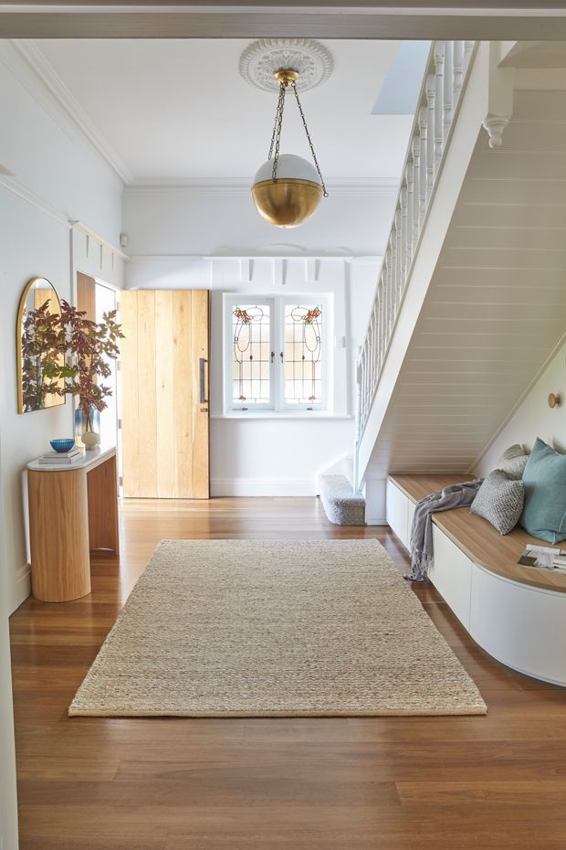 Bright, refined residential entry hall with warm timber flooring, white walls and detailed traditional trim. A stained-glass window sits beside a timber front door, while a staircase rises along the right with built-in seating and storage beneath it. The space includes a curved timber console, large neutral rug, decorative mirror, soft cushions, and a brass pendant suspended from an ornate ceiling rose.