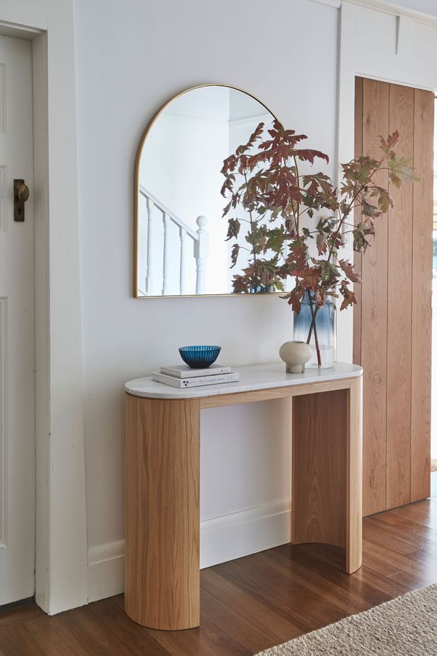A refined entryway vignette featuring a curved timber console with a pale stone top, a large arched mirror with a slim brass-toned frame, and restrained decorative objects. White painted walls, timber flooring, a concealed timber door, and a partial staircase reflection create a warm, contemporary traditional interior.
