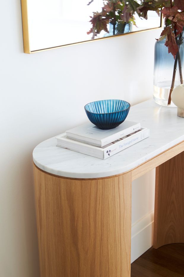 A close-up interior vignette of a curved built-in timber console with fluted or vertically grained wood cabinetry and a rounded white marble benchtop. Books and a blue decorative bowl sit on the surface, with a large gold-framed mirror and a glass vase of leafy branches above and beside it. The styling is bright, refined, and contemporary.
