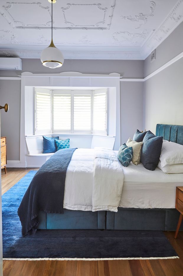 Bright, refined bedroom with a white upholstered bed dressed in layered white bedding and blue-grey throws and cushions. The room features a teal upholstered bed base and headboard, white built-in window seat cabinetry, plantation shutters, pale grey walls, timber flooring, and a dark area rug. An ornate decorative ceiling with plaster mouldings and a contemporary pendant light creates a blend of classic architectural detailing and modern furnishings.