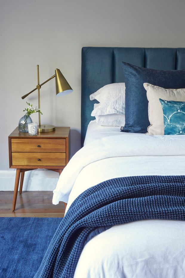 A styled bedroom featuring a bed with a tall deep-blue upholstered panelled headboard, crisp white bedding, layered navy and white cushions, and a textured navy throw. A mid-century-inspired timber bedside table with tapered legs sits beside the bed, topped with a brass articulated lamp, vase, and small decor. The room has white walls, timber flooring, and a blue area rug.