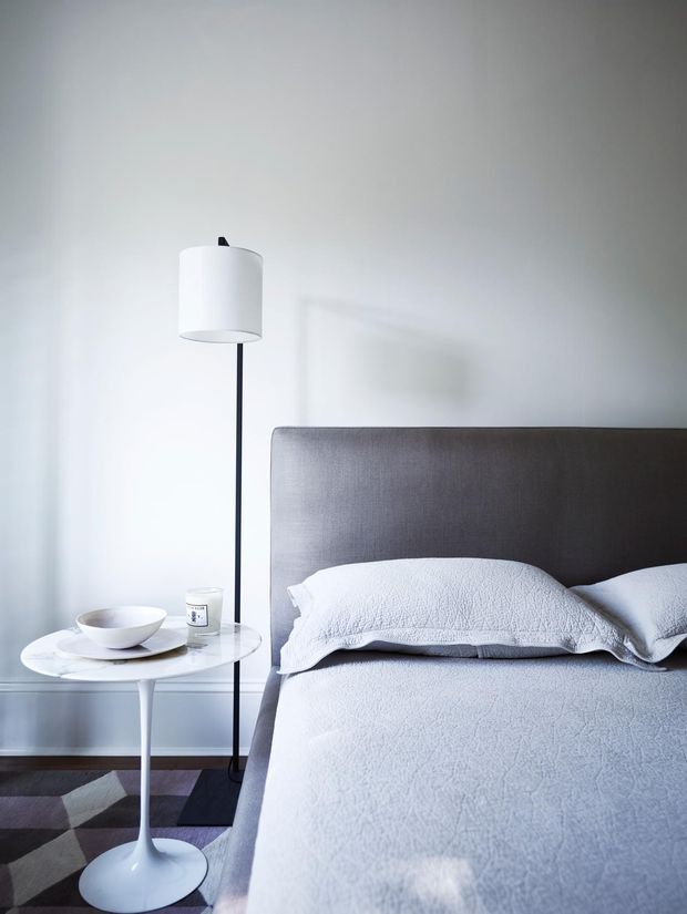 A calm, minimalist bedroom vignette featuring a large upholstered grey headboard, white textured bedding, a slim black floor lamp with a white drum shade, and a small white round pedestal side table holding a bowl and candle. The room has pale walls, dark timber flooring, and a geometric muted rug.