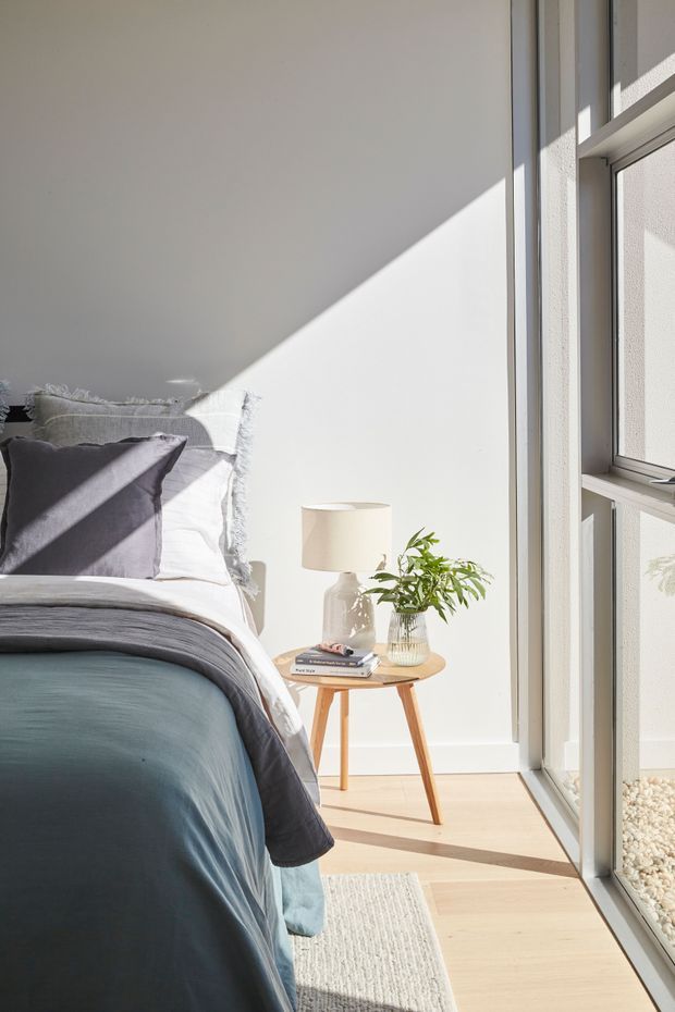 A bright, serene bedroom corner with a neatly made bed, layered grey and blue bedding, a small round timber bedside table, ceramic lamp, books, and a leafy plant. Full-height glazing brings in strong natural sunlight, casting geometric shadows across the white wall and pale timber floor. The room has a clean, contemporary and calming feel.