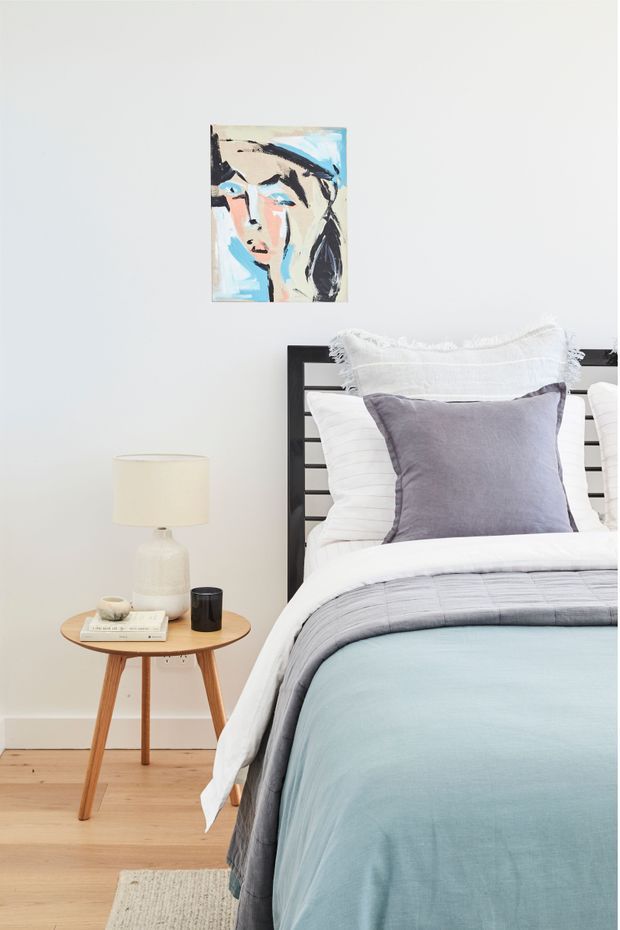 A bright, softly styled bedroom featuring a black horizontal-bar bed frame, layered white and pale blue bedding, and a muted lavender accent pillow. A small round timber side table holds a ceramic lamp, books, and a dark drinking glass, while an abstract portrait hangs above the bed. Light timber flooring and a textured neutral rug add warmth.