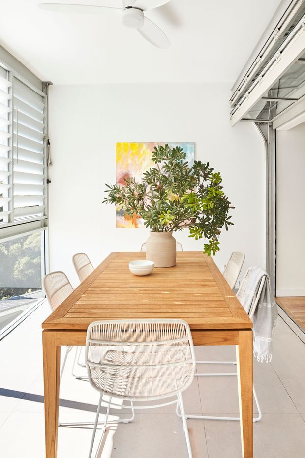 Bright dining area with a rectangular solid timber dining table, woven white dining chairs, white walls, tiled floor, plantation shutters, large glazed openings, and a ceiling fan. A large leafy plant in a ceramic vase and colourful artwork add natural and warm accents. The room is airy and sunlit with a relaxed contemporary coastal feel.
