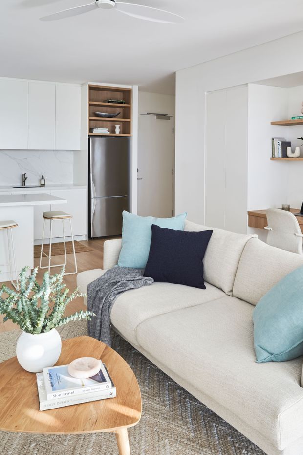 Bright open-plan apartment interior showing a compact white kitchen with marble-look splashback, integrated cabinetry and stainless-steel refrigerator, opening into a furnished living room. A cream sectional sofa with light blue and navy cushions faces a round timber coffee table, while built-in timber shelving and a small home-office nook are visible to the right. White walls, pale timber flooring, a textured rug and a white ceiling fan create an airy, contemporary setting.