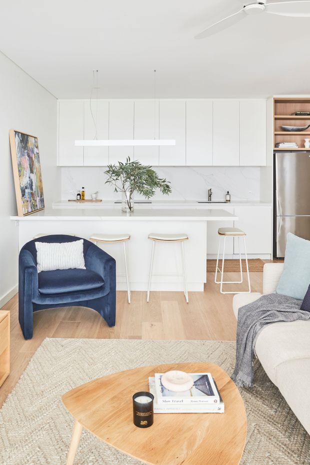 Bright open-plan living and kitchen area with white flat-panel cabinetry, a white island with slim pendant lighting, timber flooring, and a stainless-steel refrigerator. The living zone includes a navy upholstered armchair, pale sofa, textured neutral rug, and light timber coffee table. A ceiling fan and simple contemporary finishes create an airy, restrained interior.