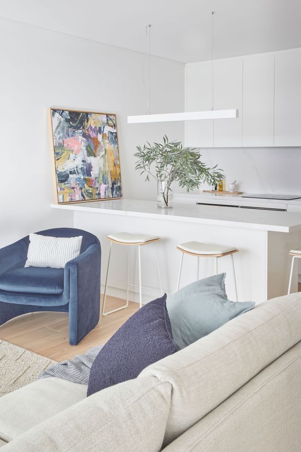 Bright open-plan living and kitchen space with white cabinetry and walls, a white island bench with integrated seating, pale timber flooring, and a linear pendant light. A deep blue armchair, neutral sofa, soft cushions, framed abstract artwork, and a leafy branch add colour and texture.