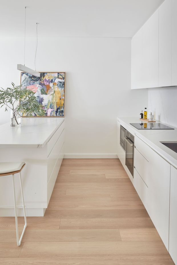 Bright, compact contemporary white kitchen with a galley-style layout and a central peninsula. The space features handleless white cabinetry, white benchtops, integrated stainless-steel appliances, pale timber flooring, slim pendant lights, and a colourful abstract artwork with a potted branch adding contrast.