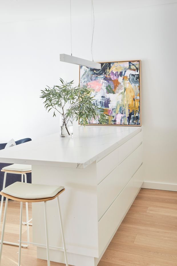 Bright, minimalist white kitchen featuring a large island with integrated handleless cabinetry, a pale timber floor, modern suspended linear lighting, bar stools, a leafy indoor plant and colourful abstract artwork.