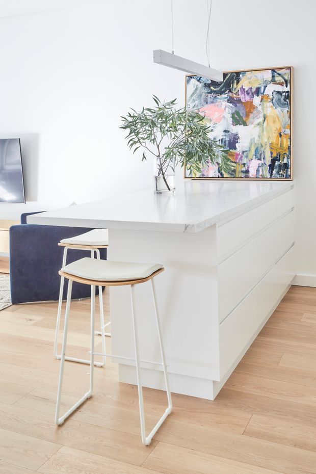 Bright contemporary kitchen interior dominated by a large white island with a smooth white benchtop and integrated white cabinetry. Two simple bar stools sit at the island, while light timber flooring, a colourful abstract artwork, a glass vase with leafy branches, and a linear suspended light add visual interest. A television is partially visible at the left edge.