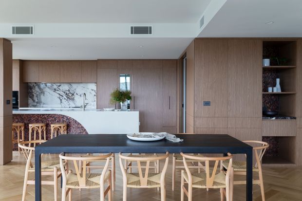 Contemporary open-plan kitchen and dining area with extensive warm timber wall panelling and cabinetry, a dark dining table surrounded by light timber dining chairs, a white stone island, marble-look splashback, and curved patterned tile detailing at the kitchen peninsula. Built-in shelving and integrated storage line the rear wall.