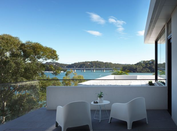 A contemporary upper-level outdoor terrace overlooking a calm waterfront inlet, with glass balustrades, pale rendered walls, dark decking, and minimalist white outdoor lounge chairs around a small round table. The terrace is connected to a modern building with extensive glazing and a broad roof overhang; a bridge and tree-covered shoreline are visible beyond.