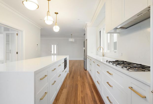 Bright, spacious contemporary white kitchen with a large central island, extensive shaker-style cabinetry, brass hardware, white benchtops, timber flooring, integrated appliances, gas cooktop, and a white tiled or slab splashback. Pendant lights and high-quality trim details create a refined, clean-lined interior.