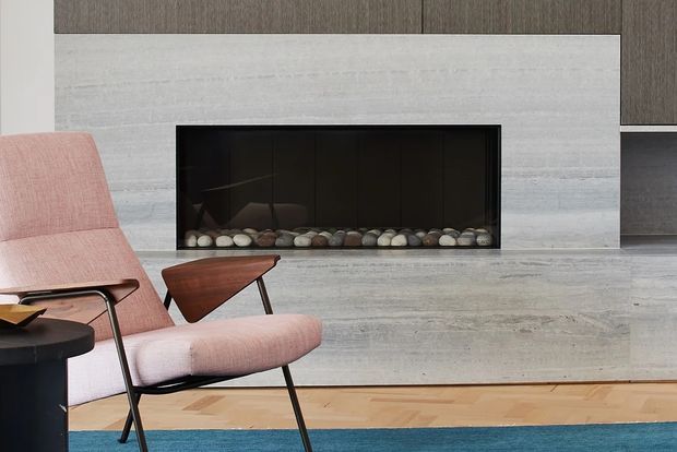 A refined modern living room centered on a wide black fireplace set into a pale stone or concrete feature wall. A pink upholstered lounge chair and sculptural timber side table sit beside a dark round table, with warm timber flooring and a blue area rug adding contrast. Dark timber cabinetry is visible above and to the right of the fireplace.