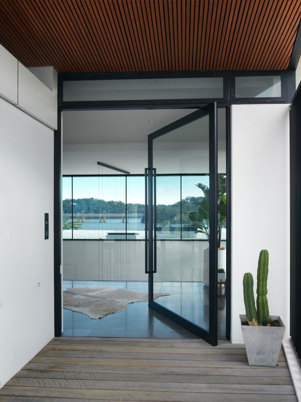 A modern entryway viewed from a timber deck, featuring a black-framed glass pivot door opening into a bright interior. The ceiling is clad in narrow warm timber battens, while white walls and dark metal framing create a crisp contemporary contrast. Through the glazing, a tiled living area, indoor plants, and a broad waterfront or lake view are visible.