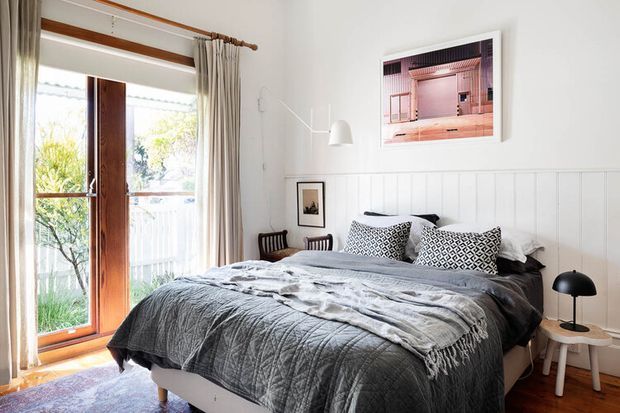 Bright, comfortably styled bedroom with a timber-framed glazed door opening to a balcony or garden. The room features white vertical wall panelling, pale walls, timber flooring, a large bed dressed in layered grey bedding, patterned cushions, a simple white wall light, and compact bedside tables. Natural light and soft neutral textiles create a relaxed, contemporary feel.