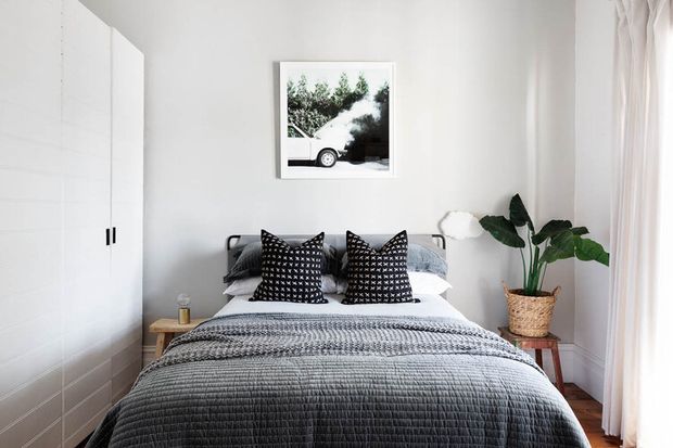 A bright, compact bedroom with a neatly made bed layered in white and grey bedding, patterned black-and-white accent cushions, white built-in wardrobes, a framed monochrome photograph, and a potted tropical plant beside sheer curtains. The room has a clean, contemporary, restrained aesthetic with warm timber flooring.