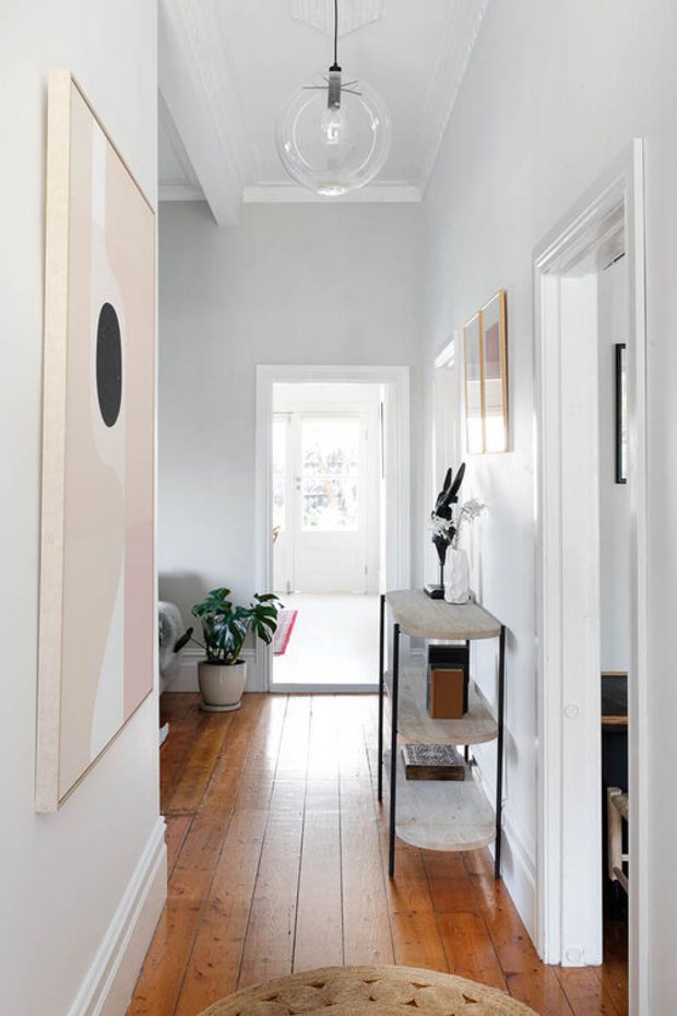 A bright, narrow residential hallway with polished warm timber floorboards, white walls and trim, and a high decorative ceiling. A slim console table with curved stone-look shelves sits along the right wall, while framed artwork, a large wall-mounted panel, a potted plant, and a glass globe pendant add visual interest. Doorways open to adjacent rooms, and daylight enters from a glazed door at the far end.
