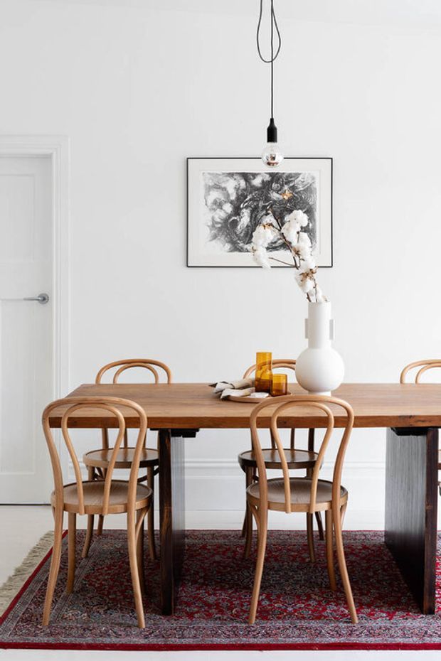 A bright, refined dining room with a long solid timber dining table surrounded by curved bentwood chairs. White walls and a white paneled door provide a clean backdrop, while a patterned red and dark rug adds contrast beneath the table. A simple black pendant light, framed artwork, amber glassware, and a white sculptural vase with dried branches complete the understated setting.