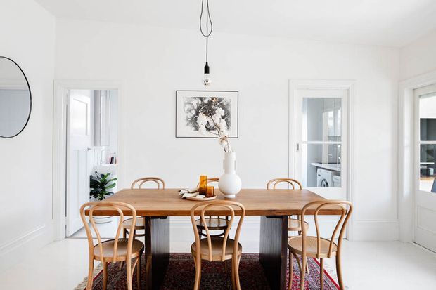 Bright, white dining room with a rectangular timber dining table surrounded by curved bentwood chairs. The room features white paneled walls and trim, tall glazed doors and windows, a simple pendant light, framed artwork, and a patterned rug beneath the table.